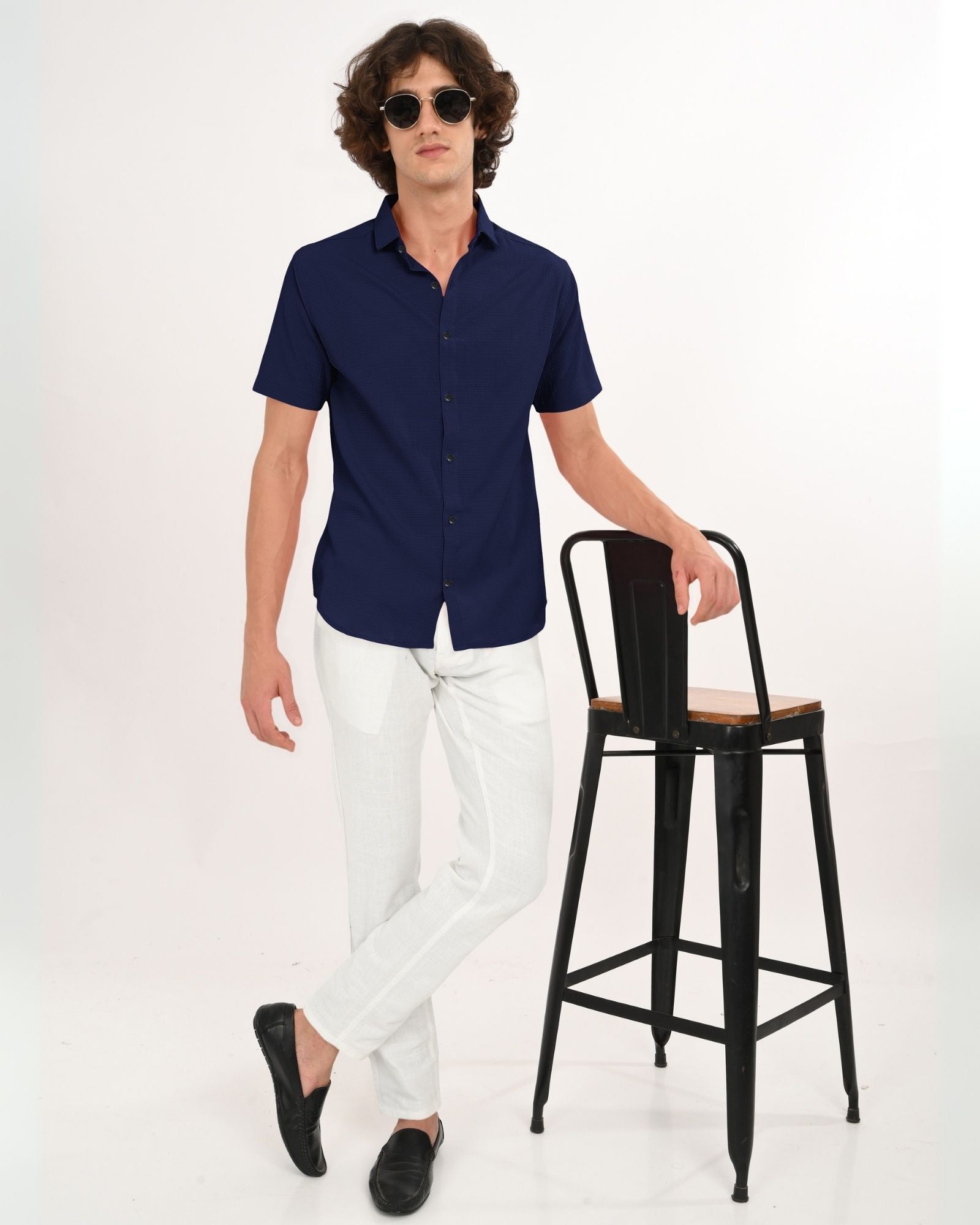 Full-body look – man wearing navy textured shirt with white pants and black loafers, standing by a chair