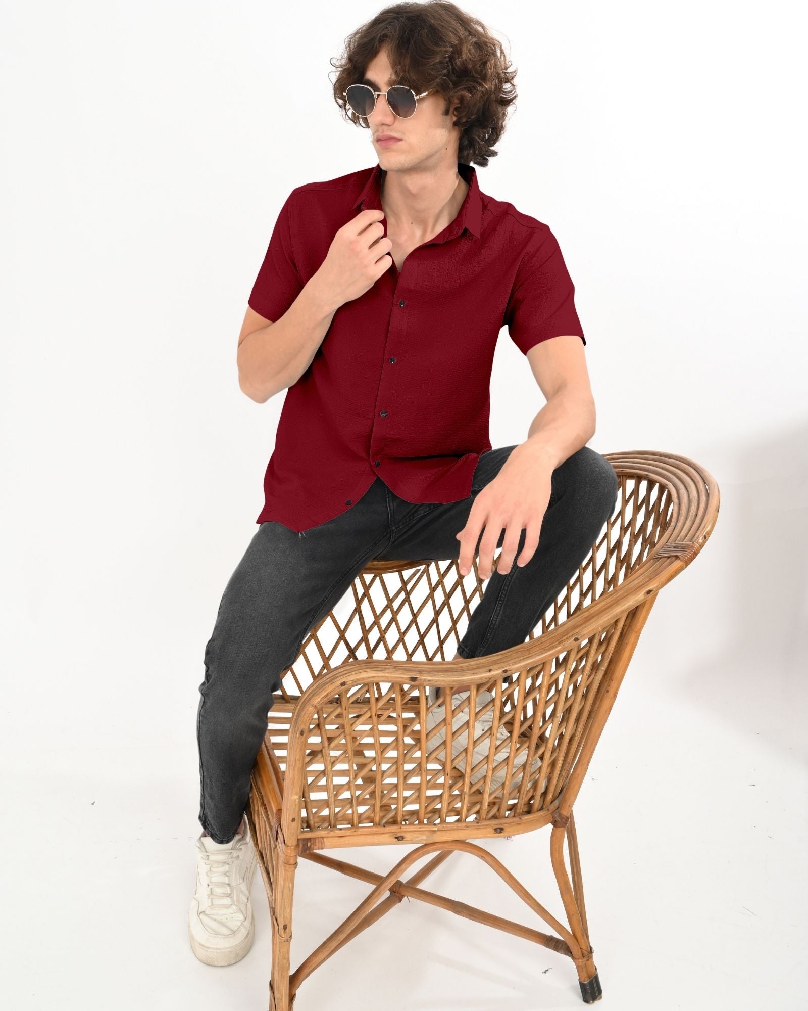 Man sitting on a wooden chair wearing a crimson red short sleeve textured shirt paired with black jeans and white sneakers.