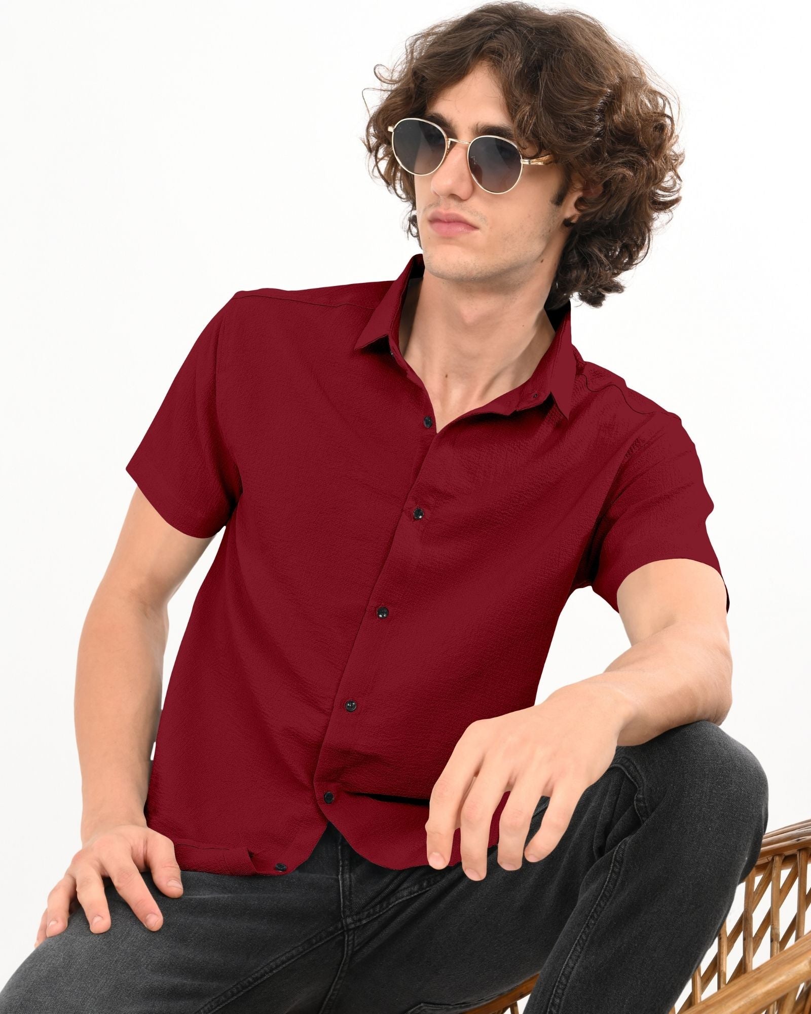 Side view of a man seated, wearing a crimson red short sleeve textured shirt paired with dark jeans.