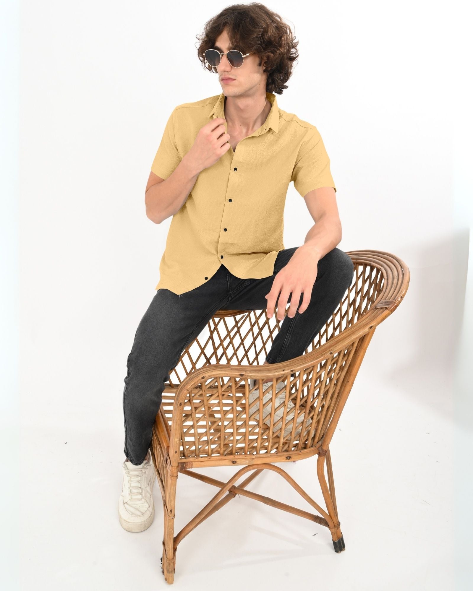 Full-body view of man in beige textured short sleeve shirt and black jeans, sitting on a wicker chair