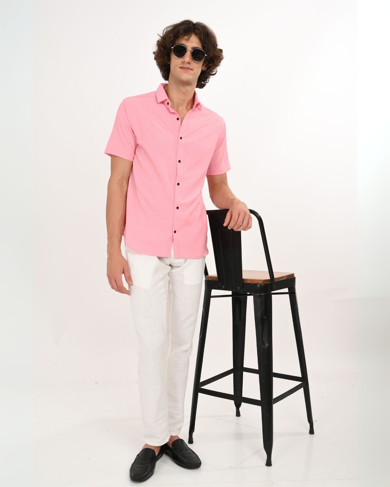 Full-length shot of man in pink button-down shirt paired with white trousers and black loafers, standing by a stool.