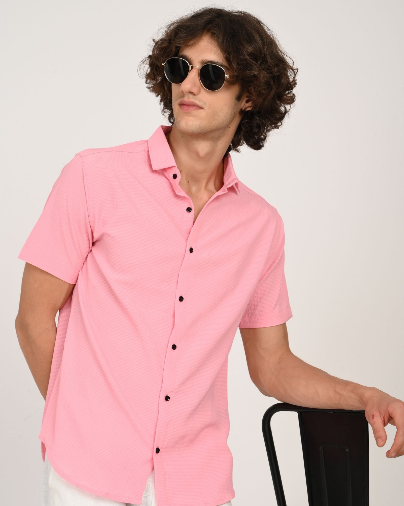 Man in pink short sleeve shirt with black buttons leaning on a stool, wearing sunglasses