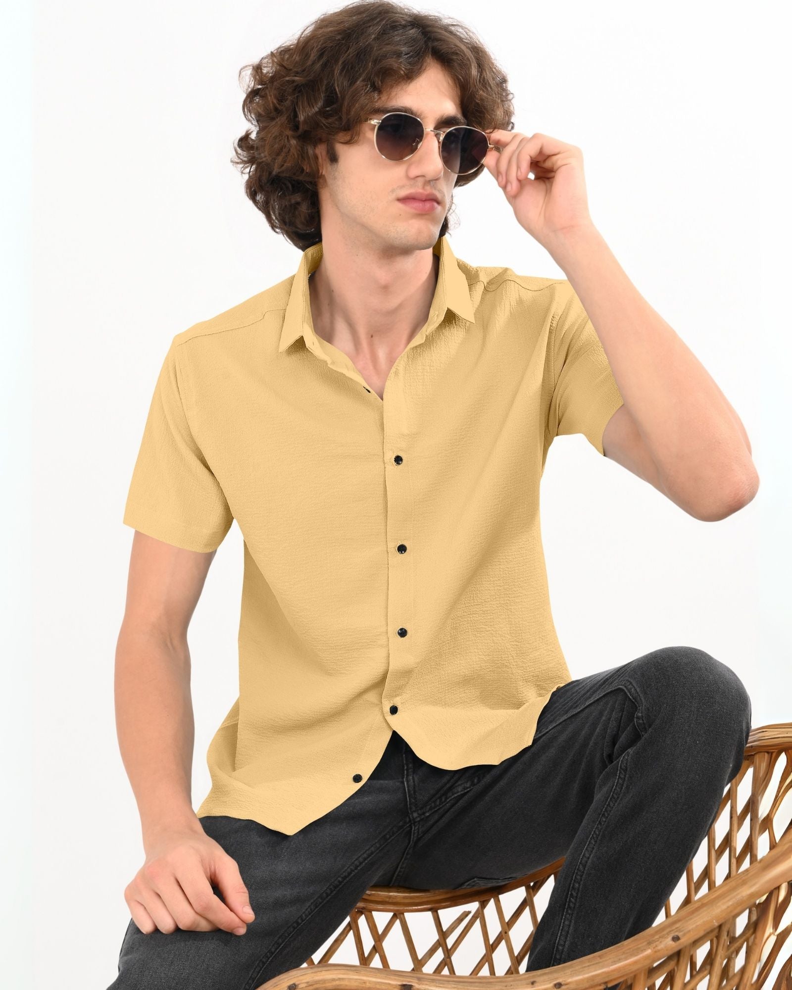 Man wearing beige textured short sleeve button-up shirt with sunglasses, seated on a chair