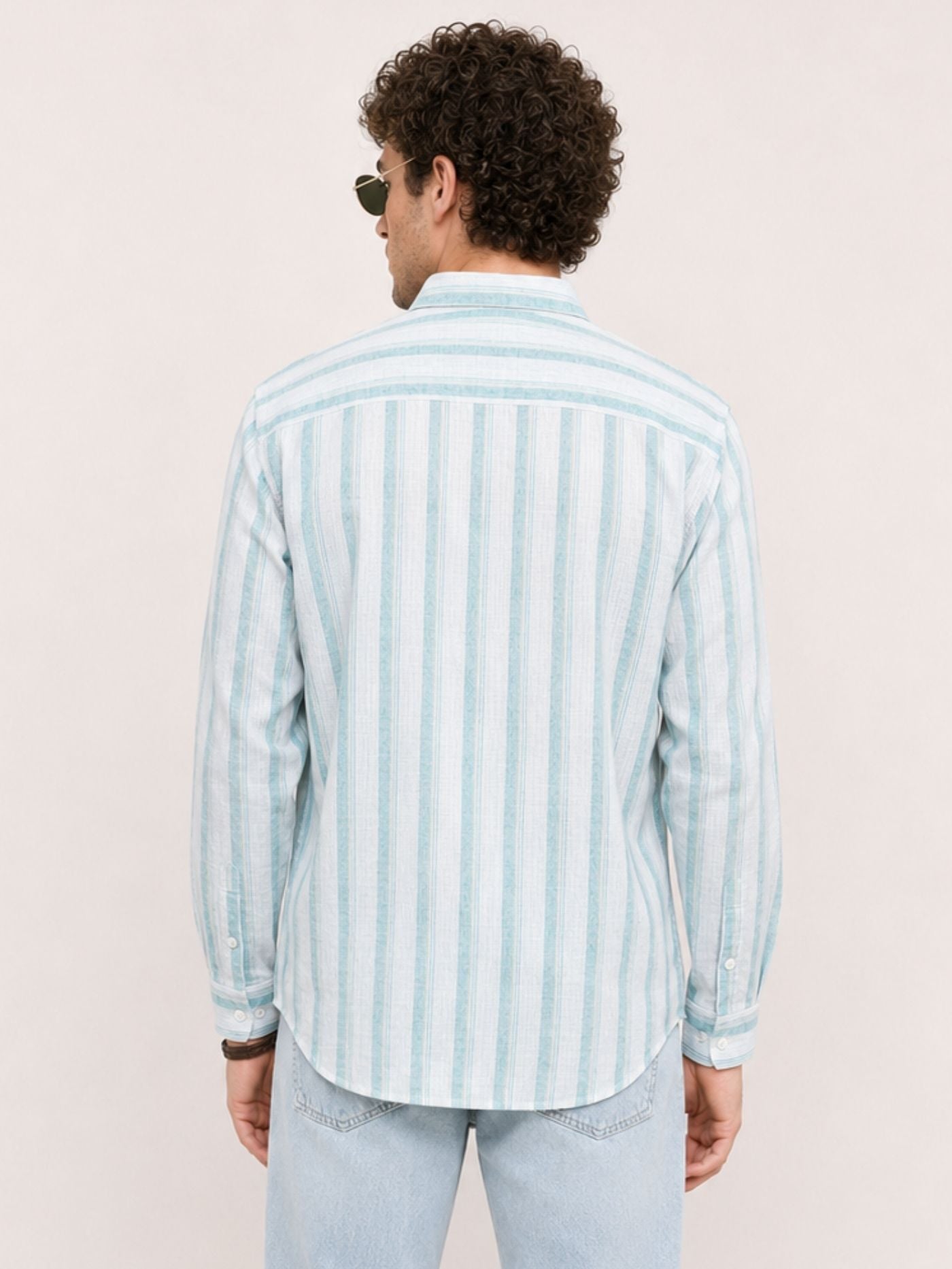 A man viewed from behind wearing a light blue and white vertically striped long-sleeve button-down shirt and light-wash jeans against a plain off-white background.