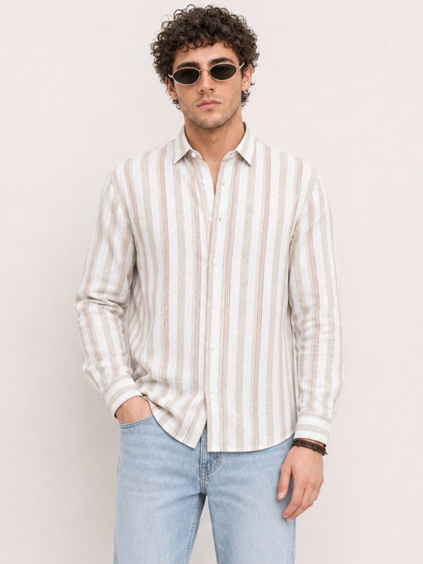 A man wearing a cream and beige vertically striped cotton button-down shirt with light blue jeans, standing against a light beige background.